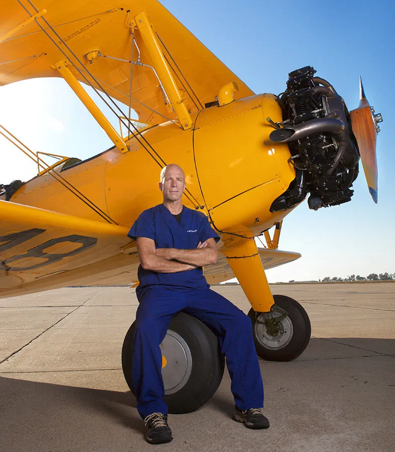 Dr. Ian Purcell in scrubs, seated beside a yellow biplane on an airfield